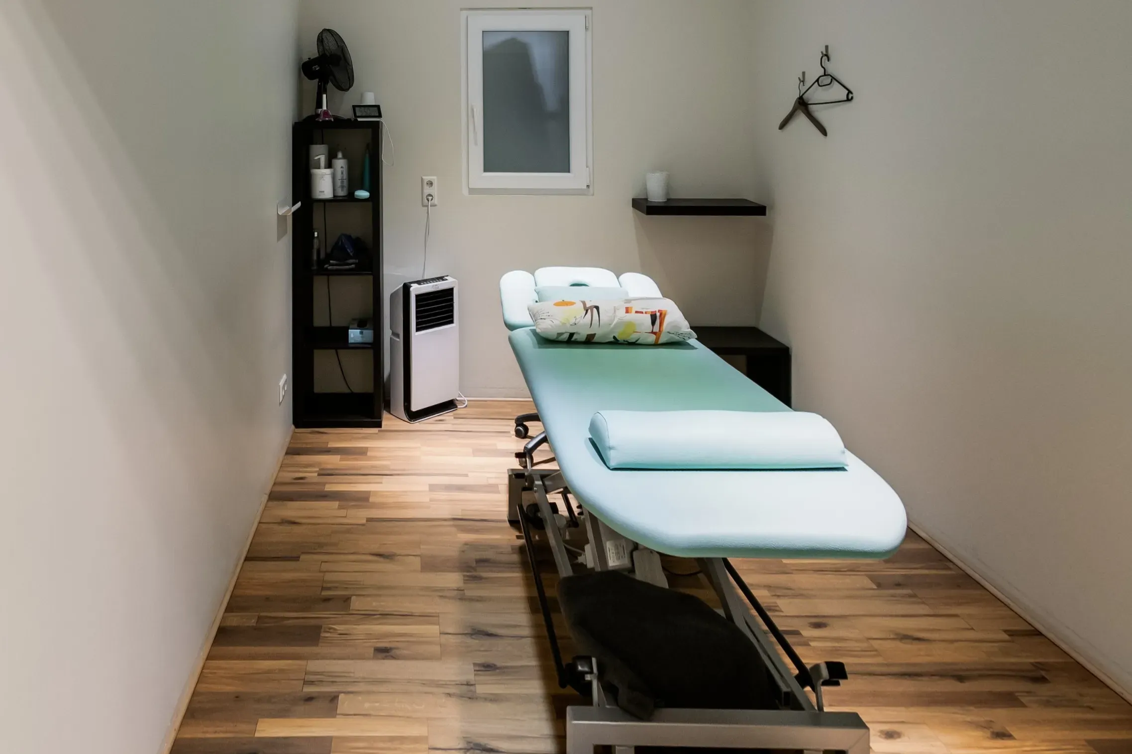 Behandlungsraum „Türkis“ mit türkisfarbener Therapieliege, Regal und Fenster